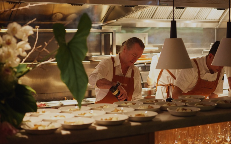 Chef Neil Perry is attending to a dish in a lit up kitchen, plates with prepared food cover the counter in front of him.
