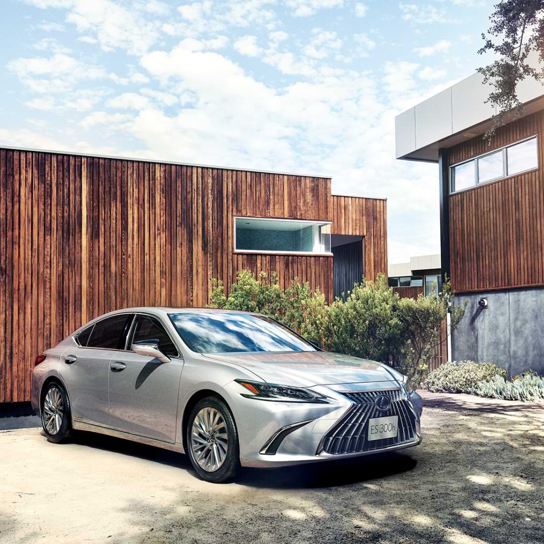 A silver ES 300h sits outside in a house's driveway, with a timber wall behind it. It is sunny.
