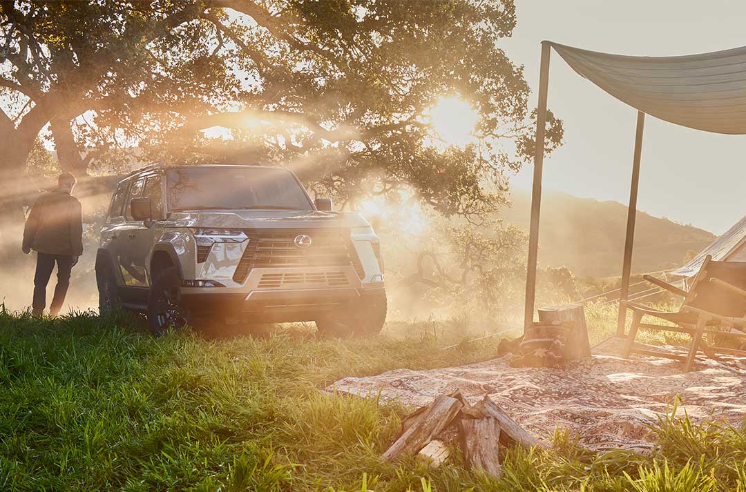 The Lexus GX is shown on grass, parked next to a campsite. The owner is shown walking beside the GX towards the campsite.