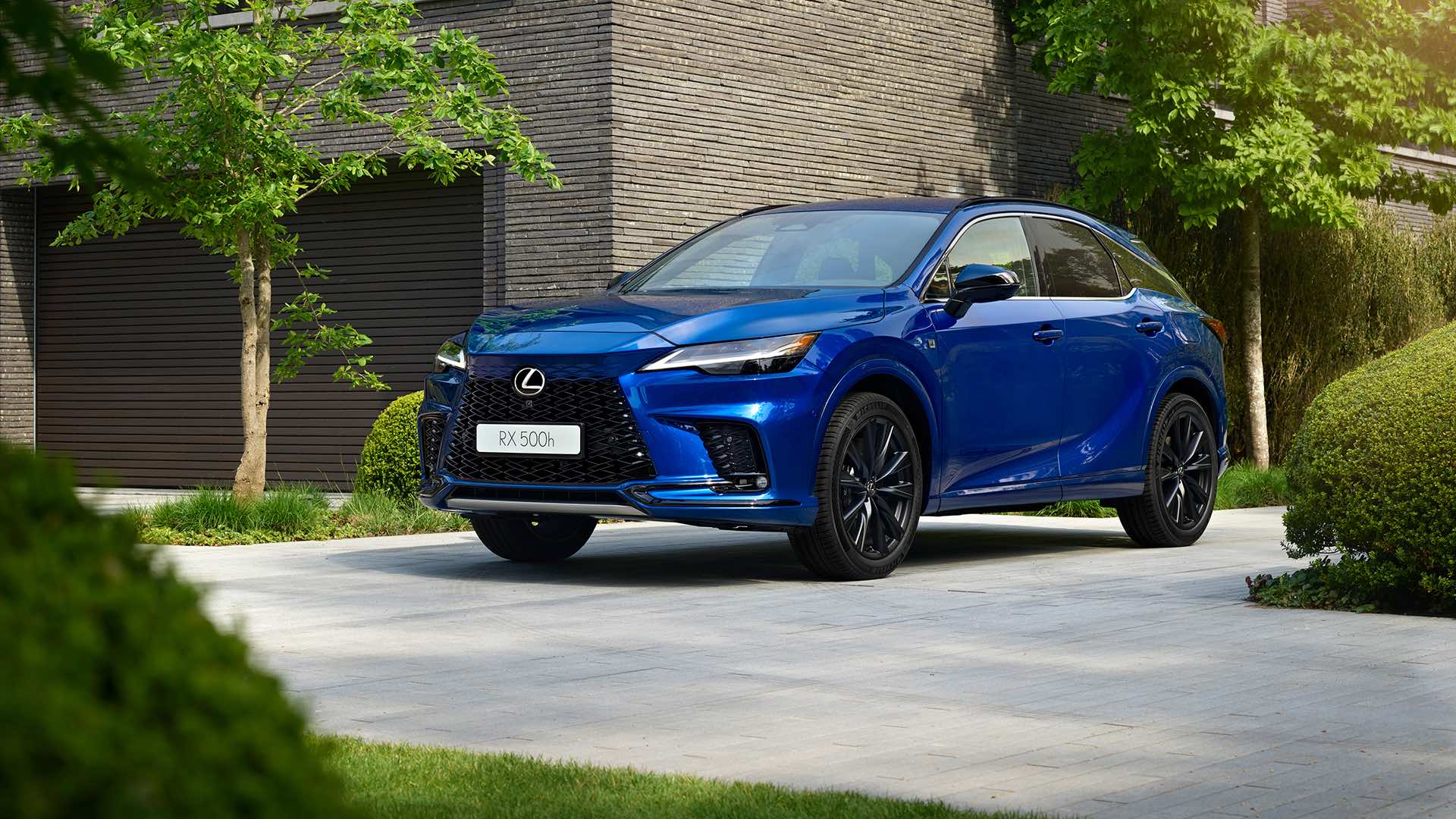 A sapphire blue Lexus RX sits on a concrete driveway. Greenery surrounds it.