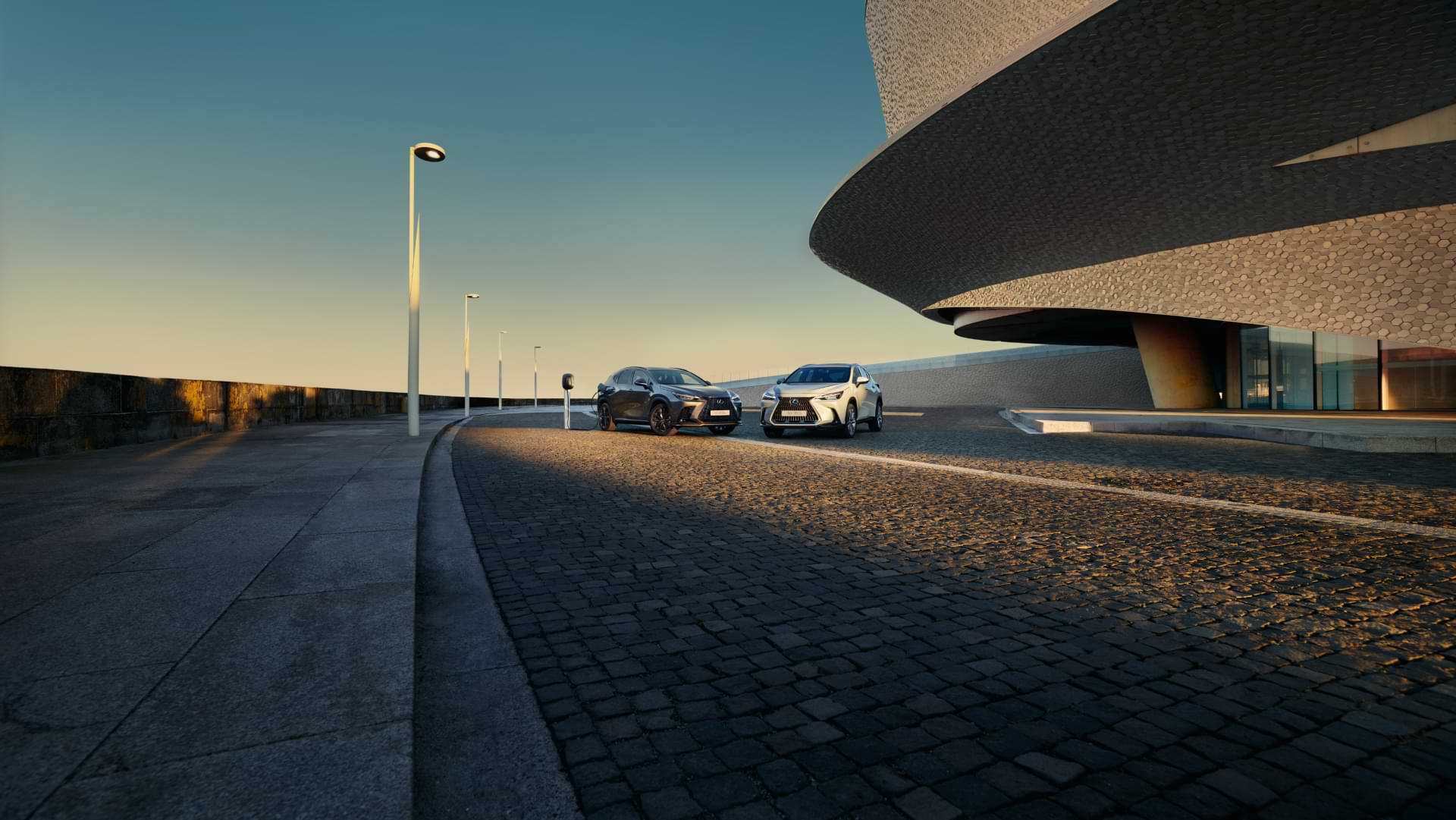 NX 450h+ and NX 350h sitting outside during the sunset. Shadows direct your eye to where they sit near a geometric building.