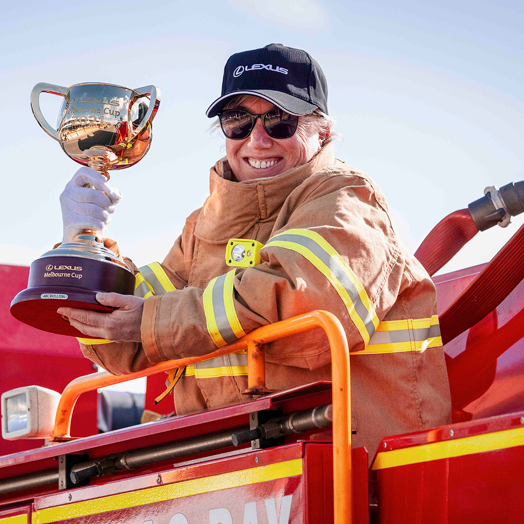 A smiling female firefighter holds the 2021 Lexus Melbourne Cup. She wears a black cap with "Lexus" written on it and stands on a red fire engine.