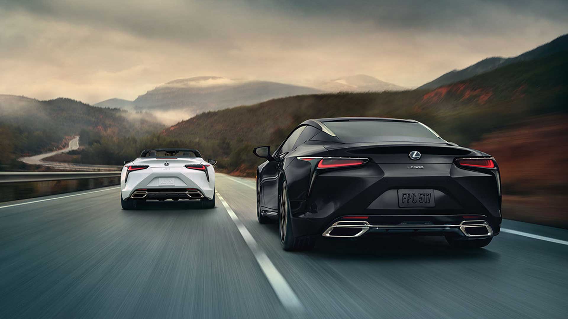 A white Lexus LC 500 Convertible drives alongside a black Lexus LC 500 coupe. They are both shown from the rear. The winding road into the mountains can be seen in the distance.