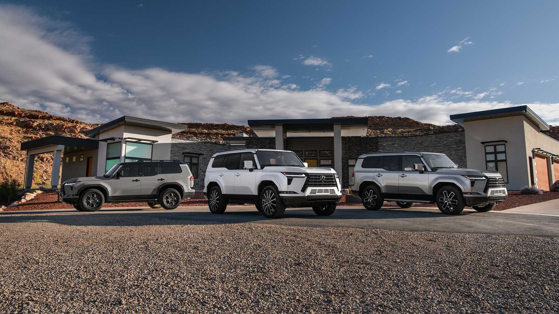 Three Lexus GX 550 SUVs, grey, white, and silver, parked in a driveway in a desert setting.