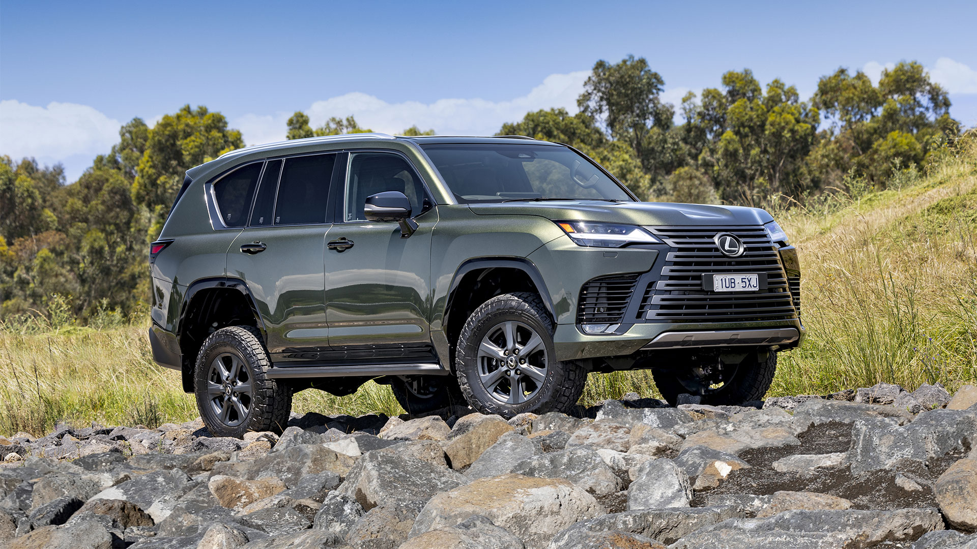 A green Lexus LX Overtrail parked on top of rocks with trees and a blue sky in the background