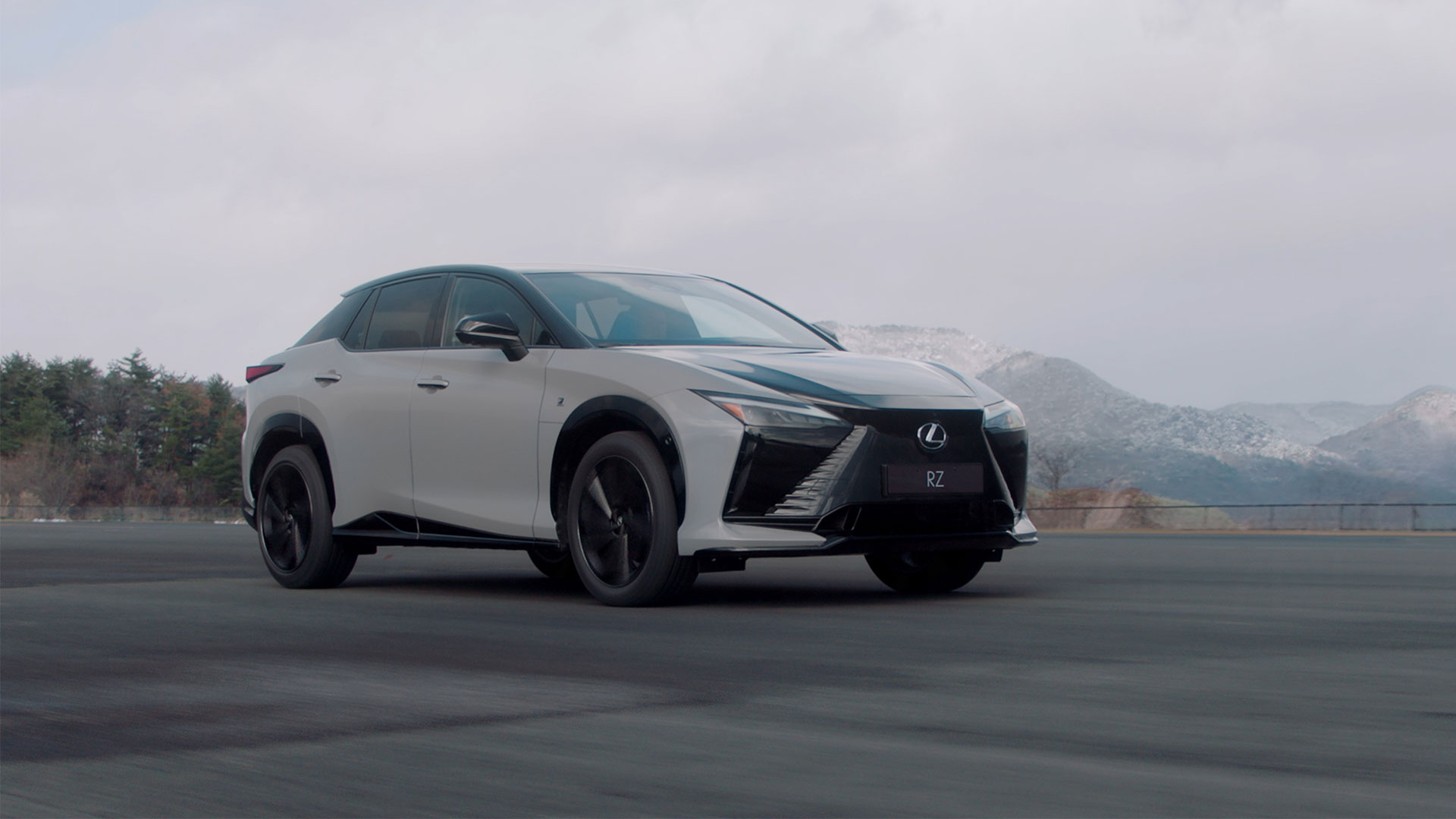 A white Lexus RZ vehicle driving on a road with mountains in the background.